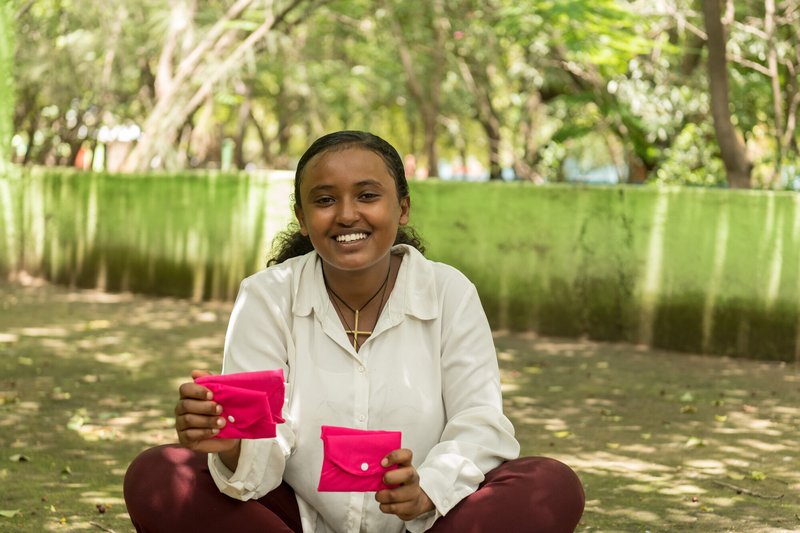 A girl sits on the ground outside holding a couple of bright pink purses (which likely contain sanitary products)