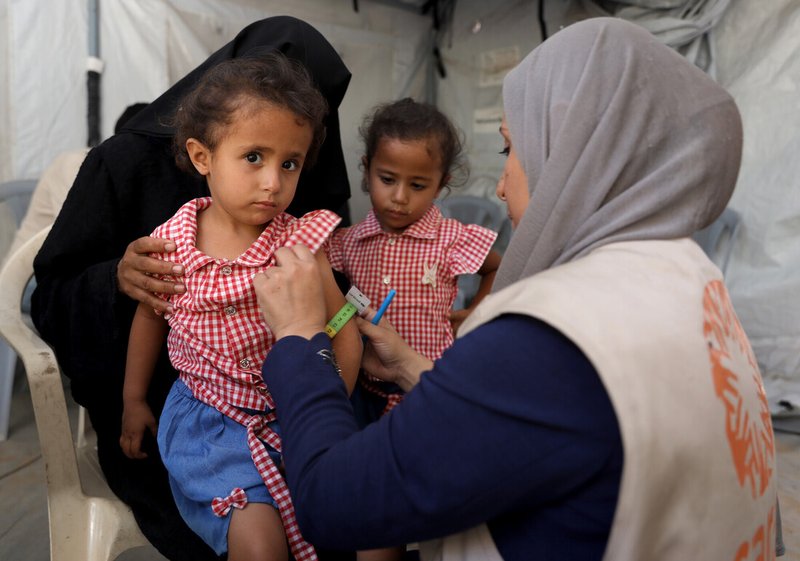 A female medic holds a measuring band around a young girl's arm. The girl is sitting on her mother's lap and another young girl looks on in the background