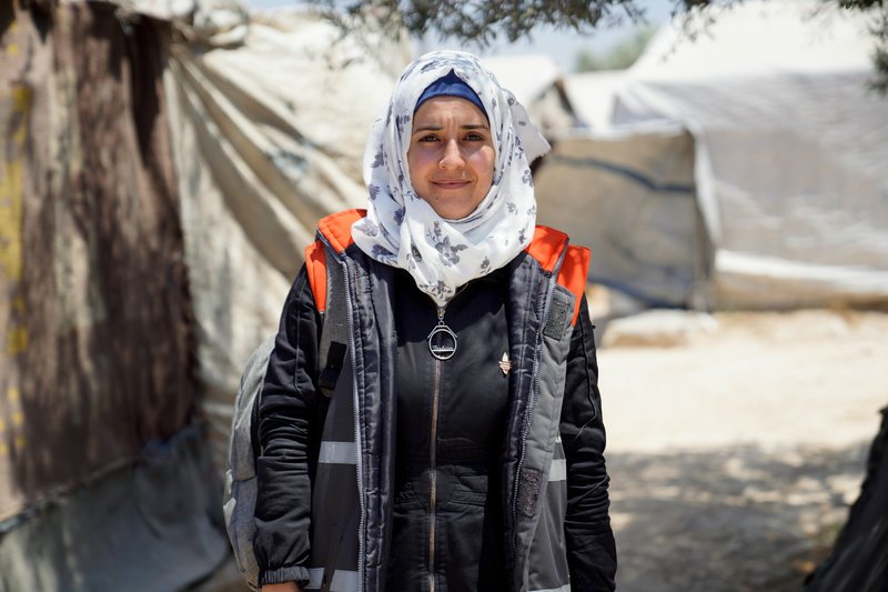 A woman wearing a headscarf smiles to camera standing in front of tents at a refugee camp
